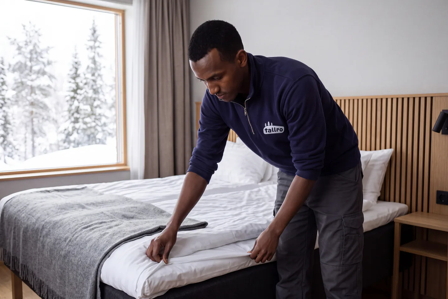 Illustration: en städare i marinblå arbetströja sträcker lakanet på en hotellsäng med snöig skog utanför fönstret.