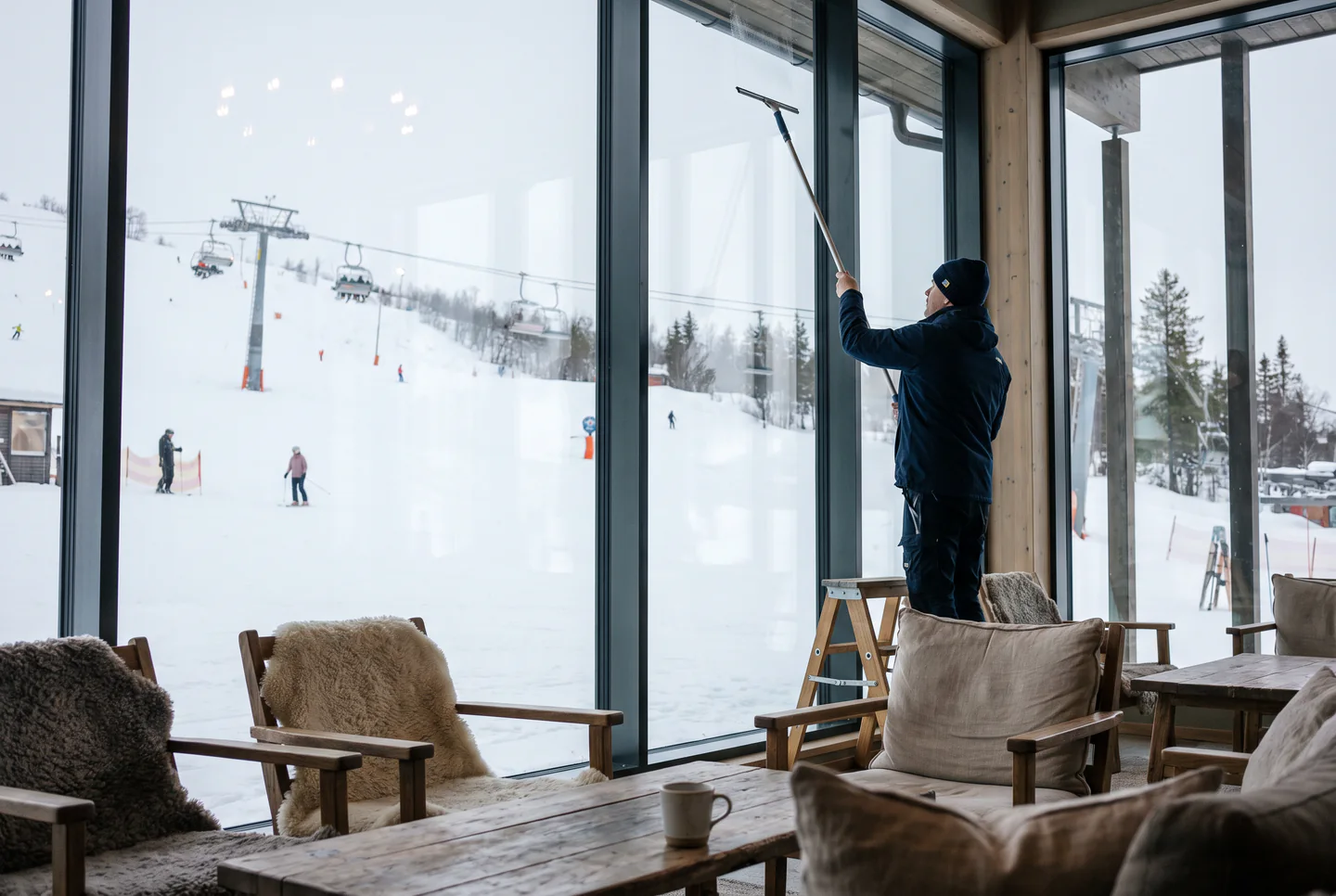 Illustration: en fönsterputsare tvättar den höga glasväggen i en hotellobby med utsikt över skidbacken.
